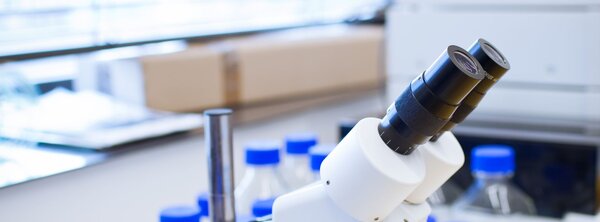 Microscope on a lab bench surrounded by clear plastic reagent bottles with blue caps in a bright laboratory with window blinds.