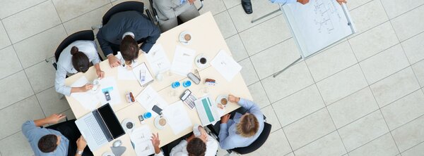Top‑down view of a group of business people sitting around a table in a meeting, with papers, laptops, coffee cups, and one person standing at a whiteboard.