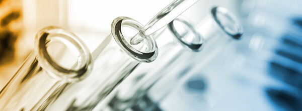 Close‑up of glass test tubes lined up with a dropper pipette above them, in a scientific laboratory setting with soft blue and orange lighting.